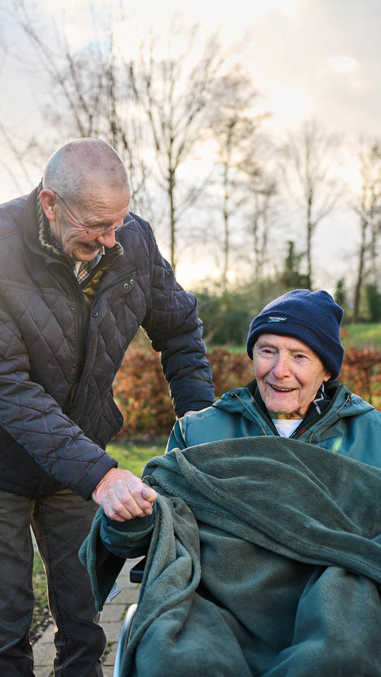 Twee mannen zijn buiten. 1 man helpt lachend een andere man die in roelstoel zit met dekentje om hem heen, de man in rolstoel lacht ook De kracht van de Anna Zorggroep zit in de kleinschalige, goede en brede basiszorg.